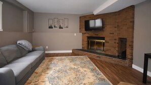 A living room with a gray sofa, a patterned rug, a brick fireplace, and a flat-screen TV mounted above it. - Benbow Suite (Hinton)