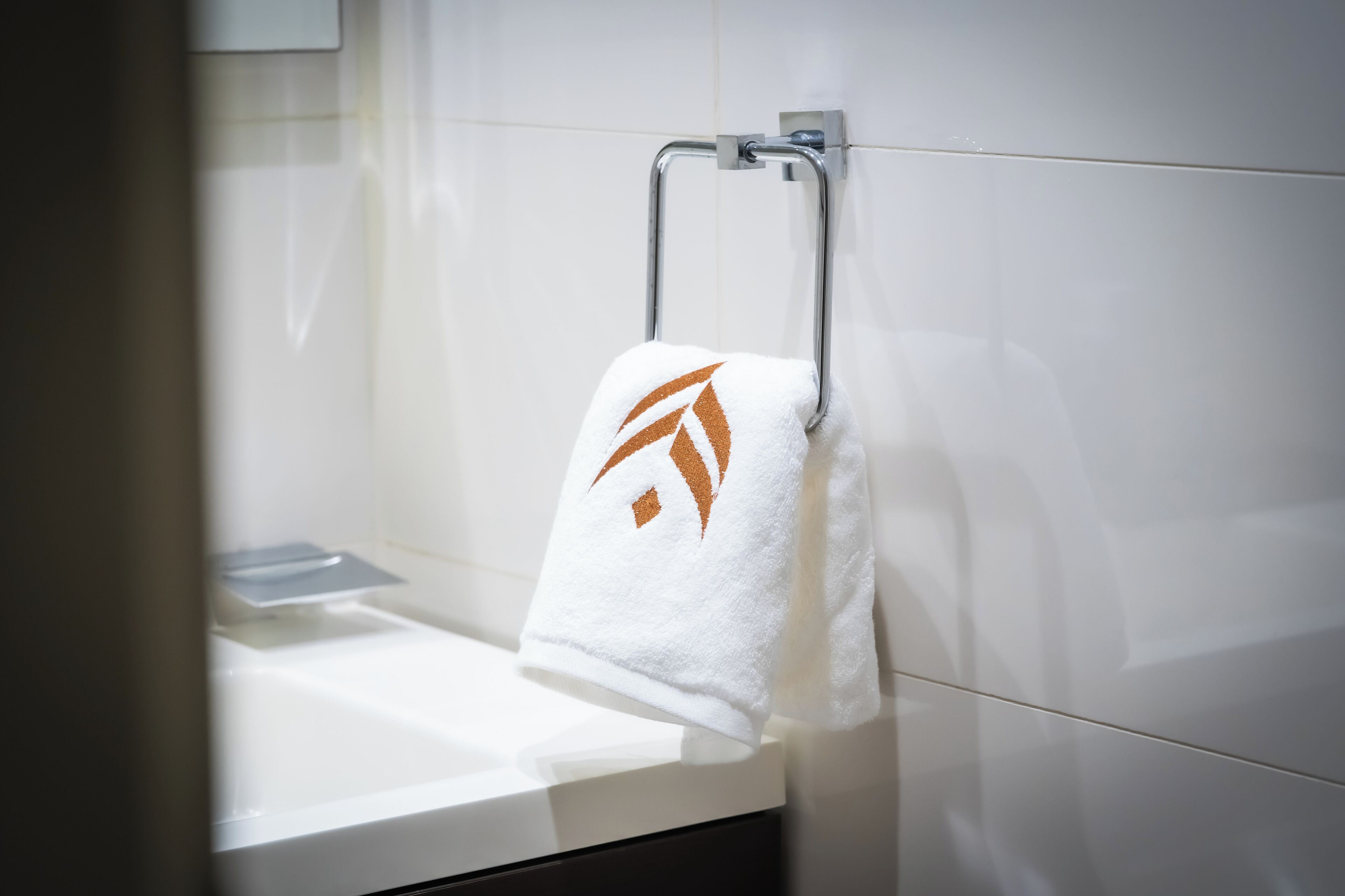 Una toalla blanca con un logo marrón colgada en un perchero metálico en un baño con azulejos blancos.