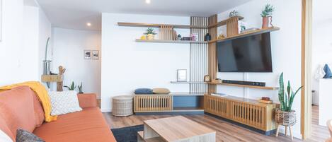 Una sala de estar moderna con sofá, mesa de centro y televisor. Se observan estantes con plantas y libros.