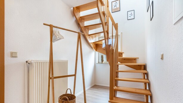 Una escalera en espiral de madera en una habitación con paredes blancas, un radiador y una cesta sobre un soporte de madera.