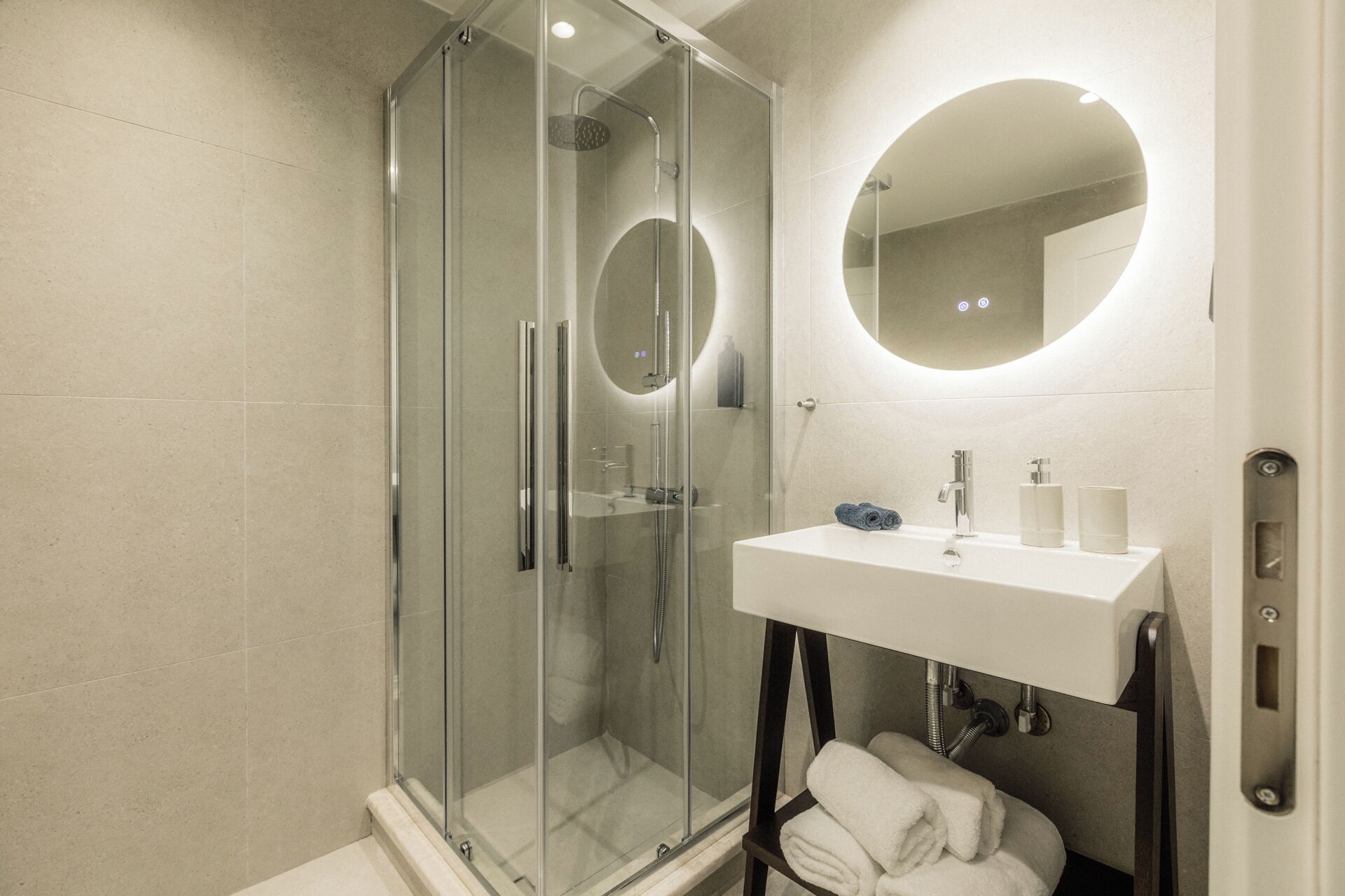 Un baño moderno con ducha envuelta en vidrio, un lavamanos blanco y un espejo redondo, y toallas dobladas con cuidado.
