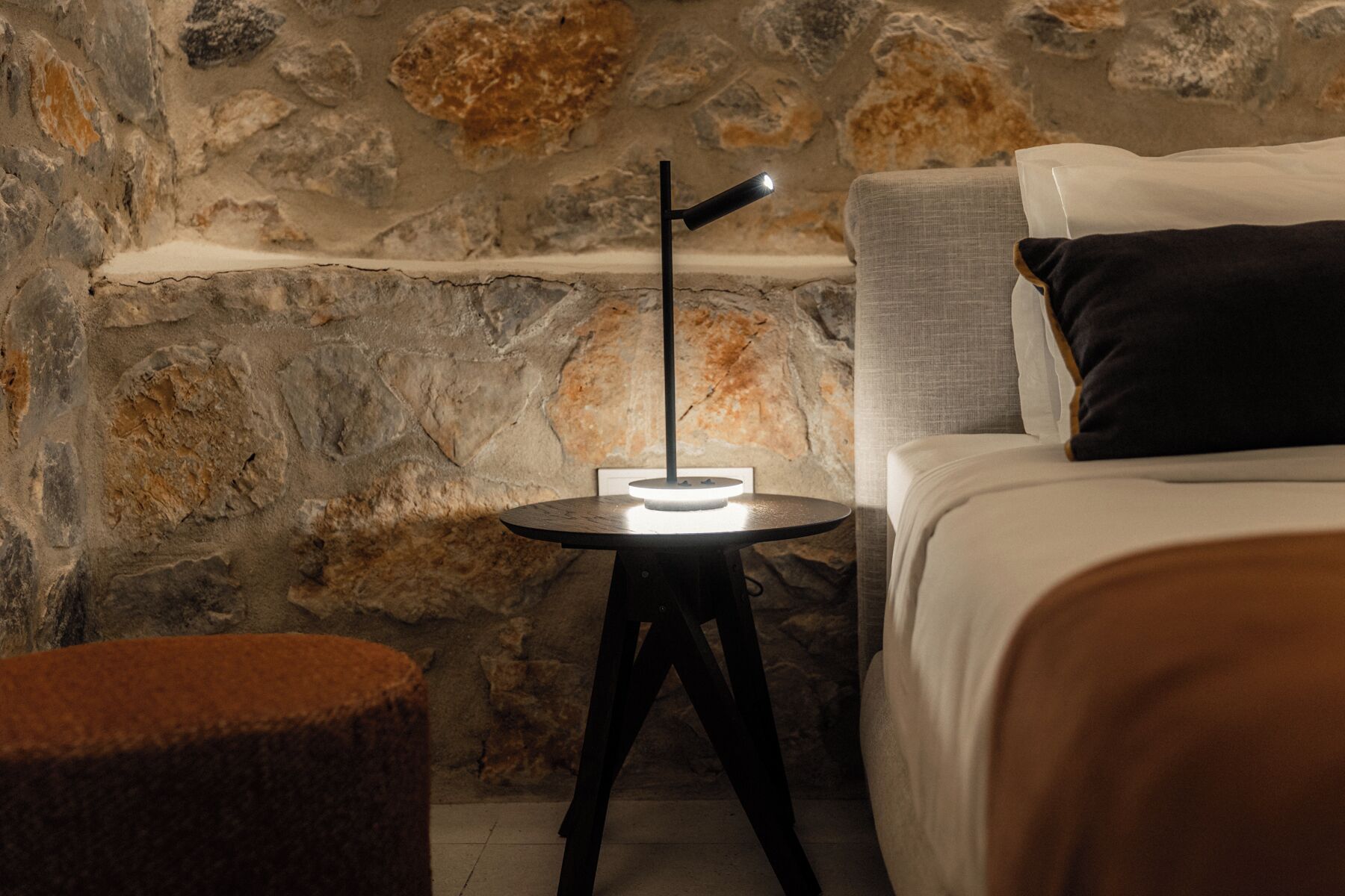 Una pared de piedra, una mesa redonda con una lámpara, una cama con almohadas y un pequeño reposapiés.