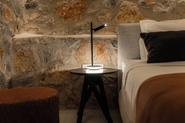 Una pared de piedra, una mesa redonda con una lámpara, una cama con almohadas y un pequeño reposapiés.
