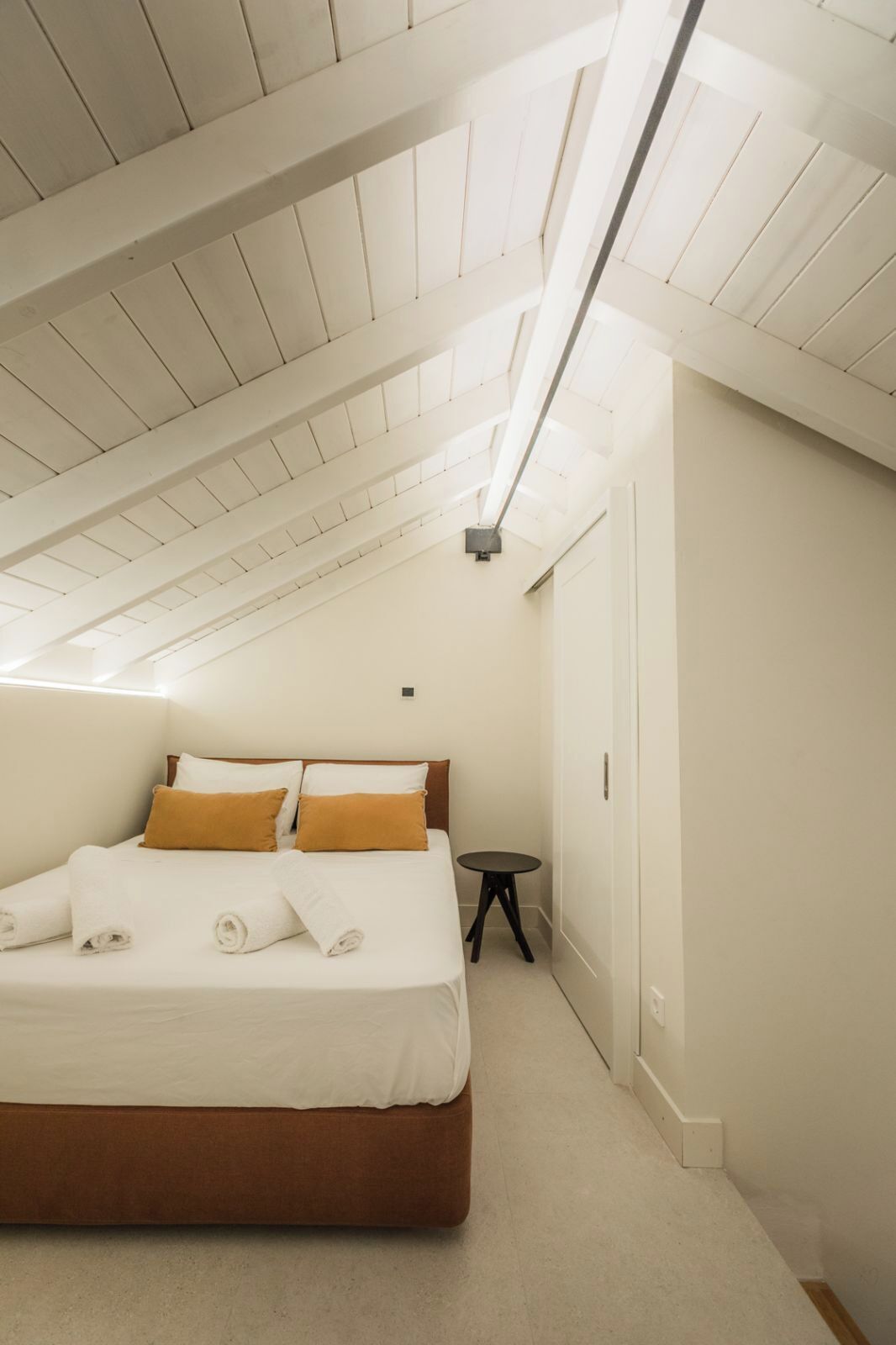 Ein Schlafzimmer mit schrägen Wänden, einem Bett mit weißer Bettwäsche, einem kleinen runden Tisch und einer Tür.