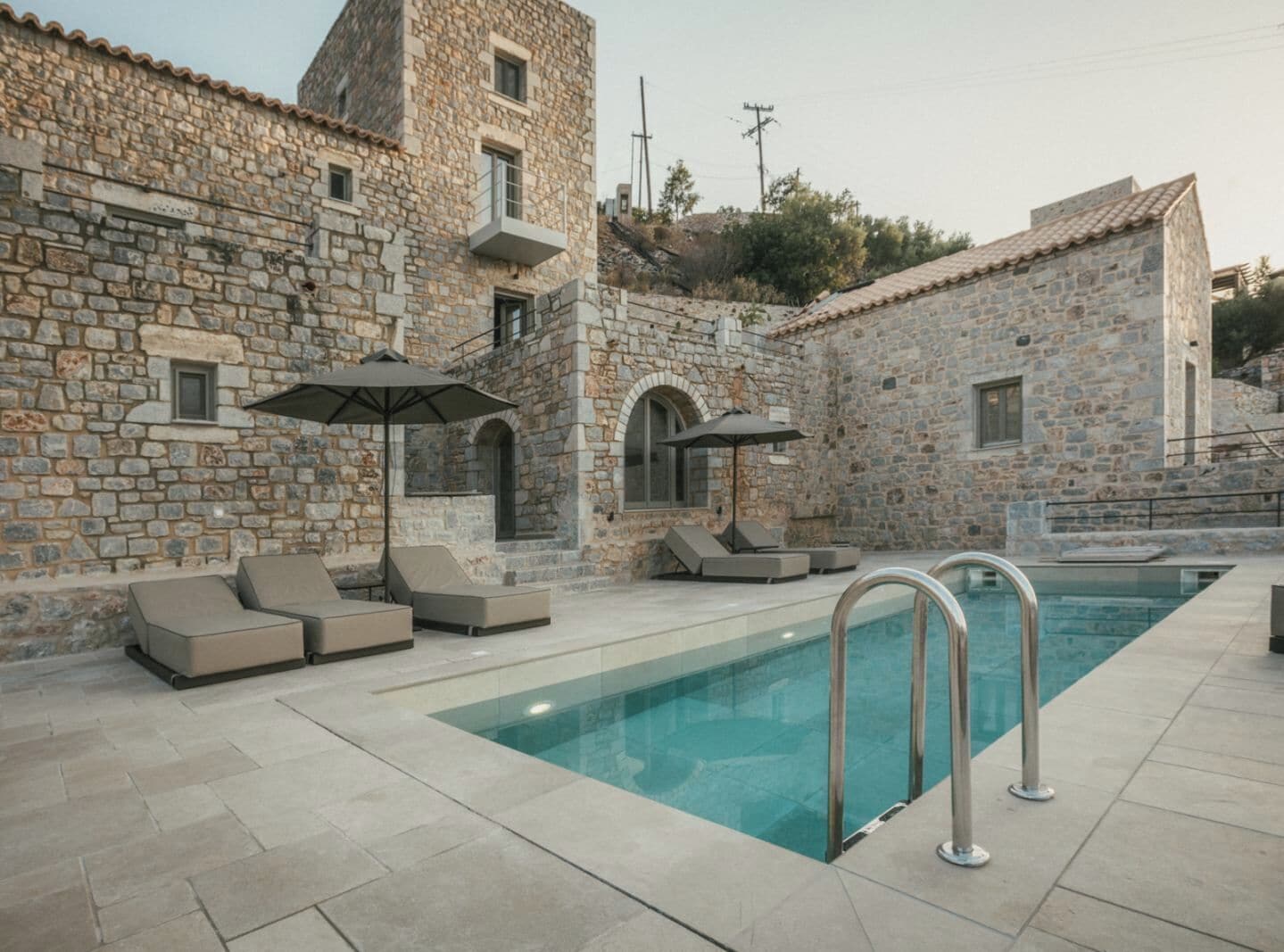 A stone building with a swimming pool, lounge chairs, and umbrellas.