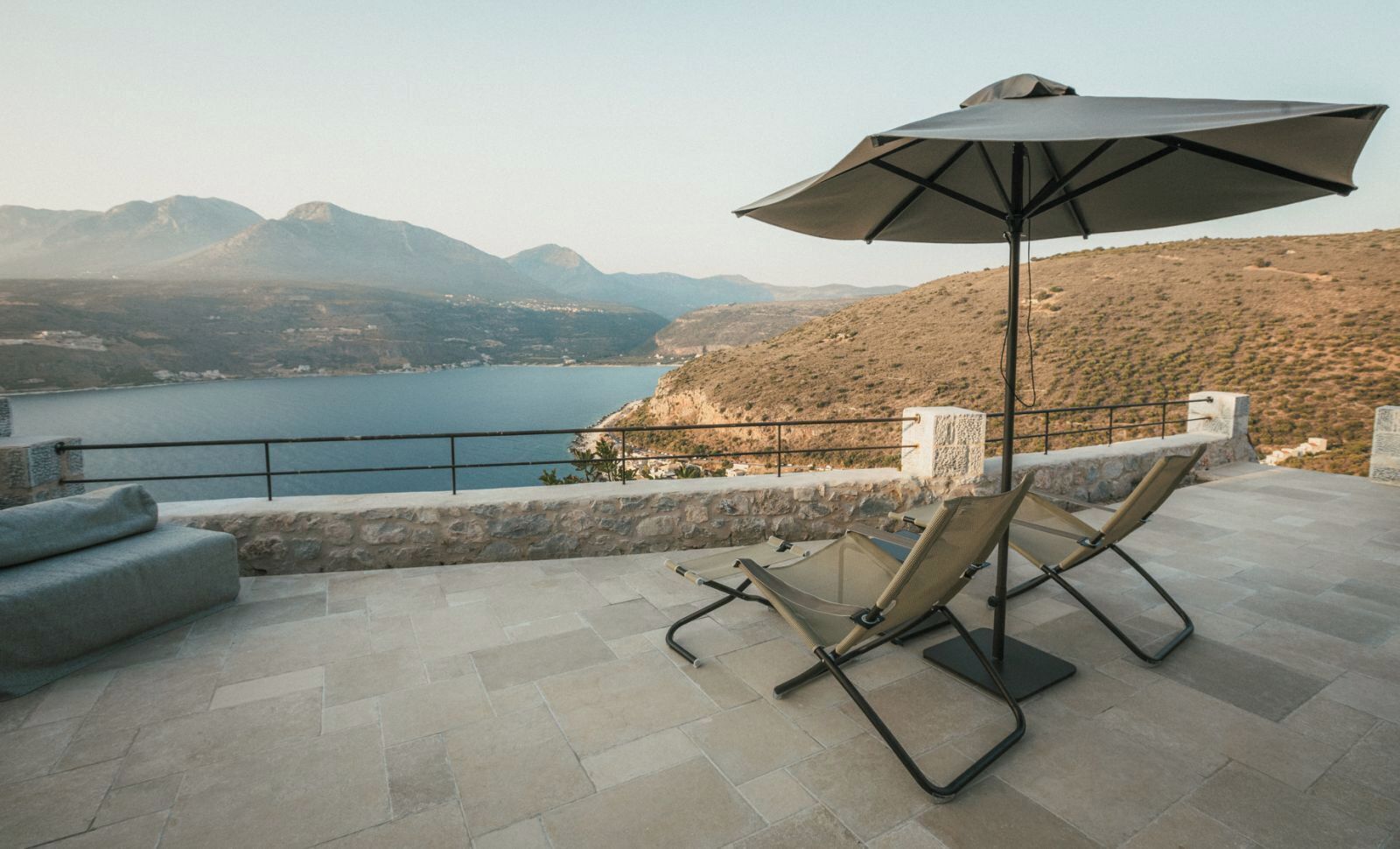 Eine Terrasse mit zwei Liegestühlen und einem Sonnenschirm, mit Blick auf ein Gewässer und Hügel.