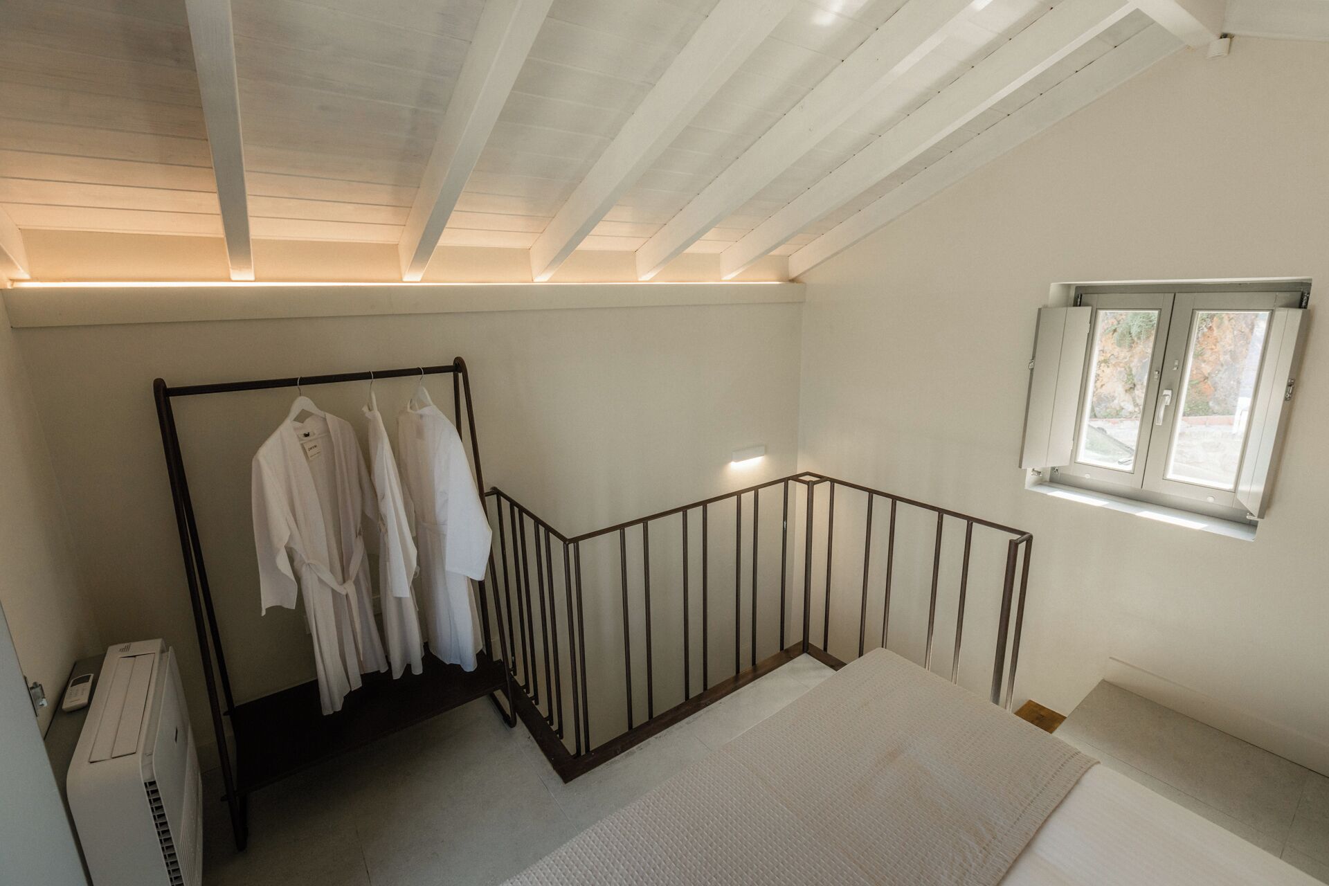 Een kamer met een schuine kap, een raam, een metalen hekwerk en een kledingrek met witte kleding.