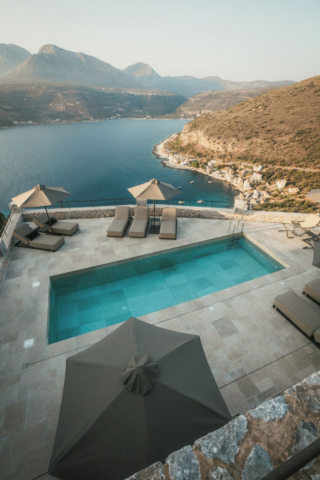 Piscina com espreguiçadeiras e guarda-sóis, com vista para a costa e montanhas ao fundo.