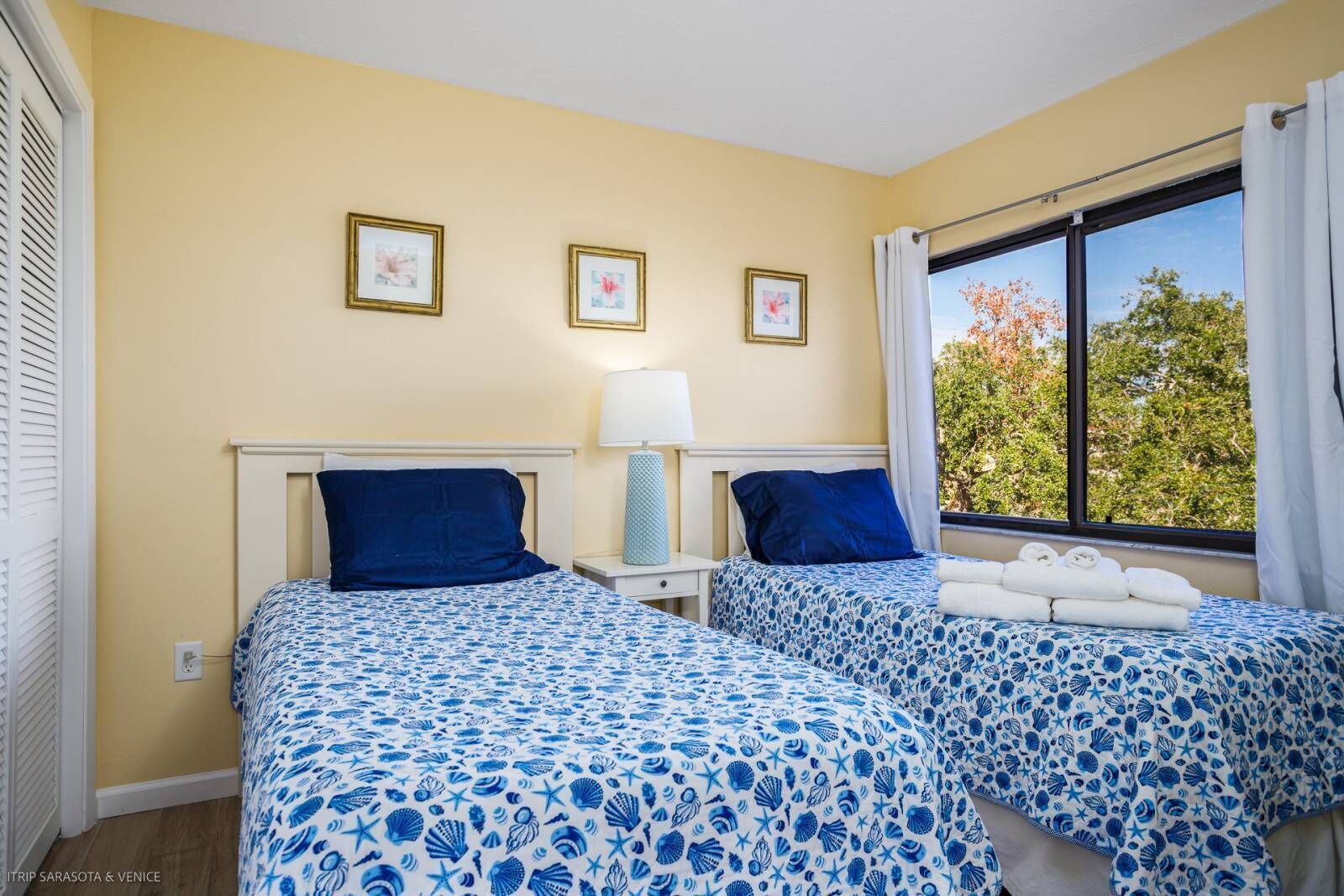 A room with two beds, each with a blue and white patterned bedspread, a bedside table with a lamp, and a window with a view of trees.