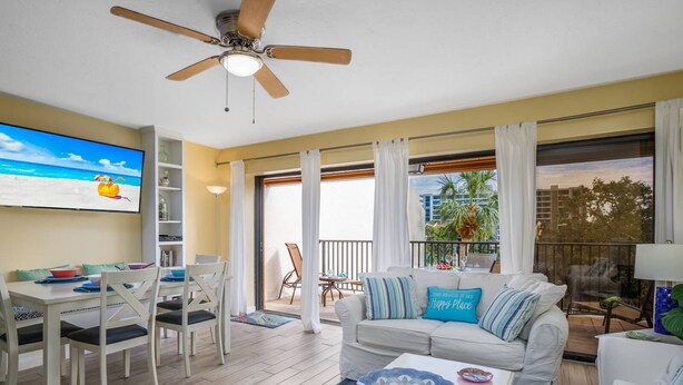 Una sala de estar con ventilador de techo, un televisor mostrando una escena de playa, un comedor con sillas y una mesa, un sofá con almohadones decorativos, y un balcón con vista a palmeras.