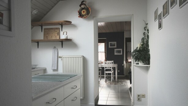 A kitchen with white cabinets, a countertop, and a hanging decorative item. There is a doorway leading to a dining area with a table and chairs. - Cozy 2-room apartment in the southern Black Forest (Steinen)