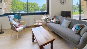 Un salon moderne avec un mur en pierre, une grande fenêtre donnant sur les arbres, un canapé gris agrémenté de coussins à motifs, une table basse en bois et un lampadaire.