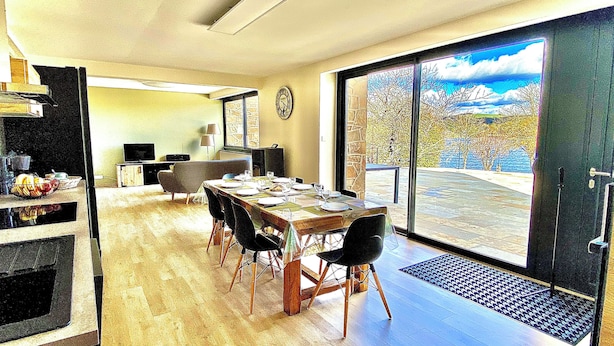 Un coin repas avec une table dressée pour six personnes, une horloge au mur et une vue sur un paysage montagneux, visible à travers de grandes portes coulissantes.