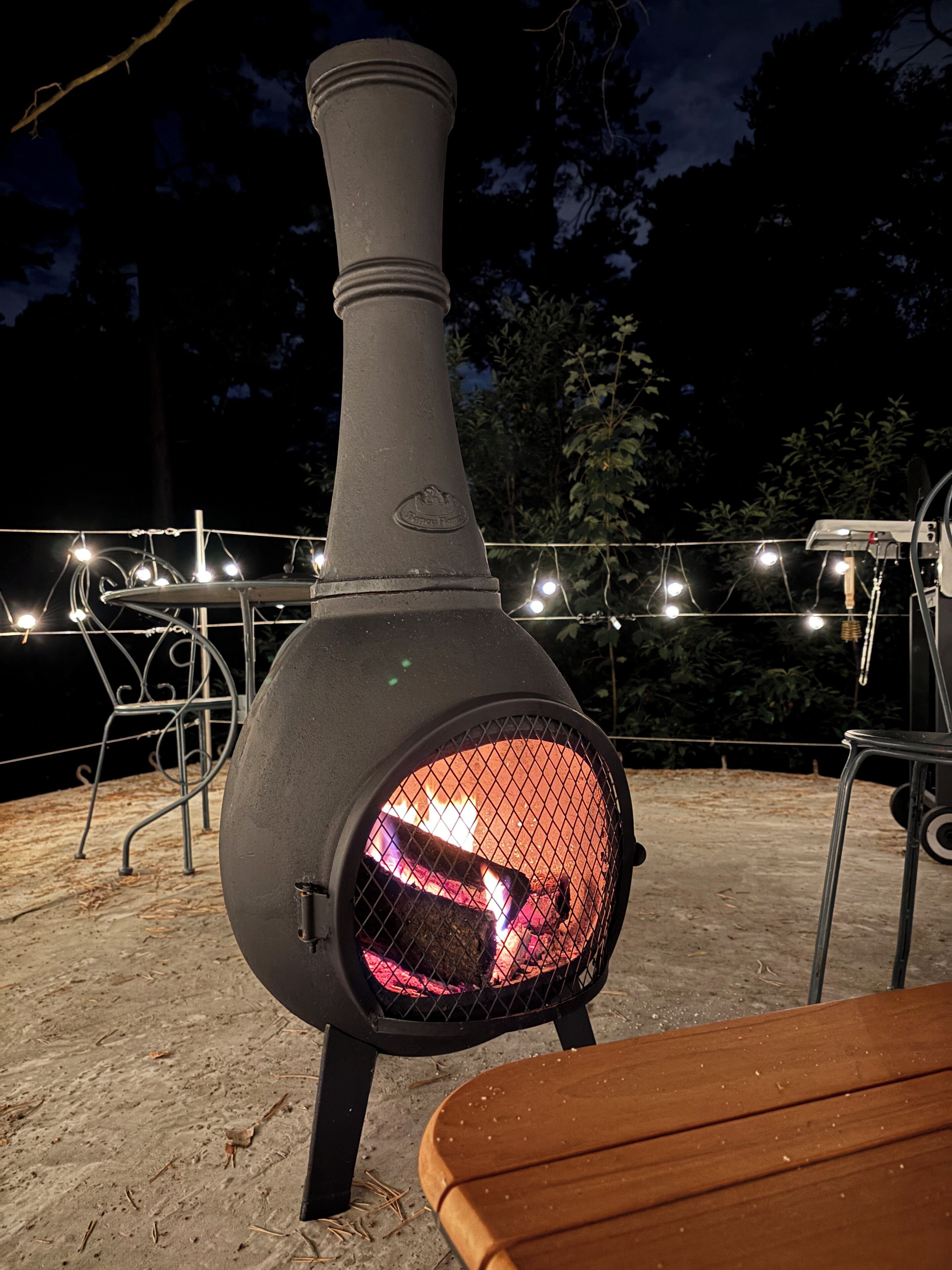 Un horno de pizza de leña con chimenea, rodeado de mobiliario exterior y luces colgantes.