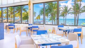 Ein Restaurant mit gedeckten Tischen, großen Fenstern und Blick auf Palmen sowie das Meer.