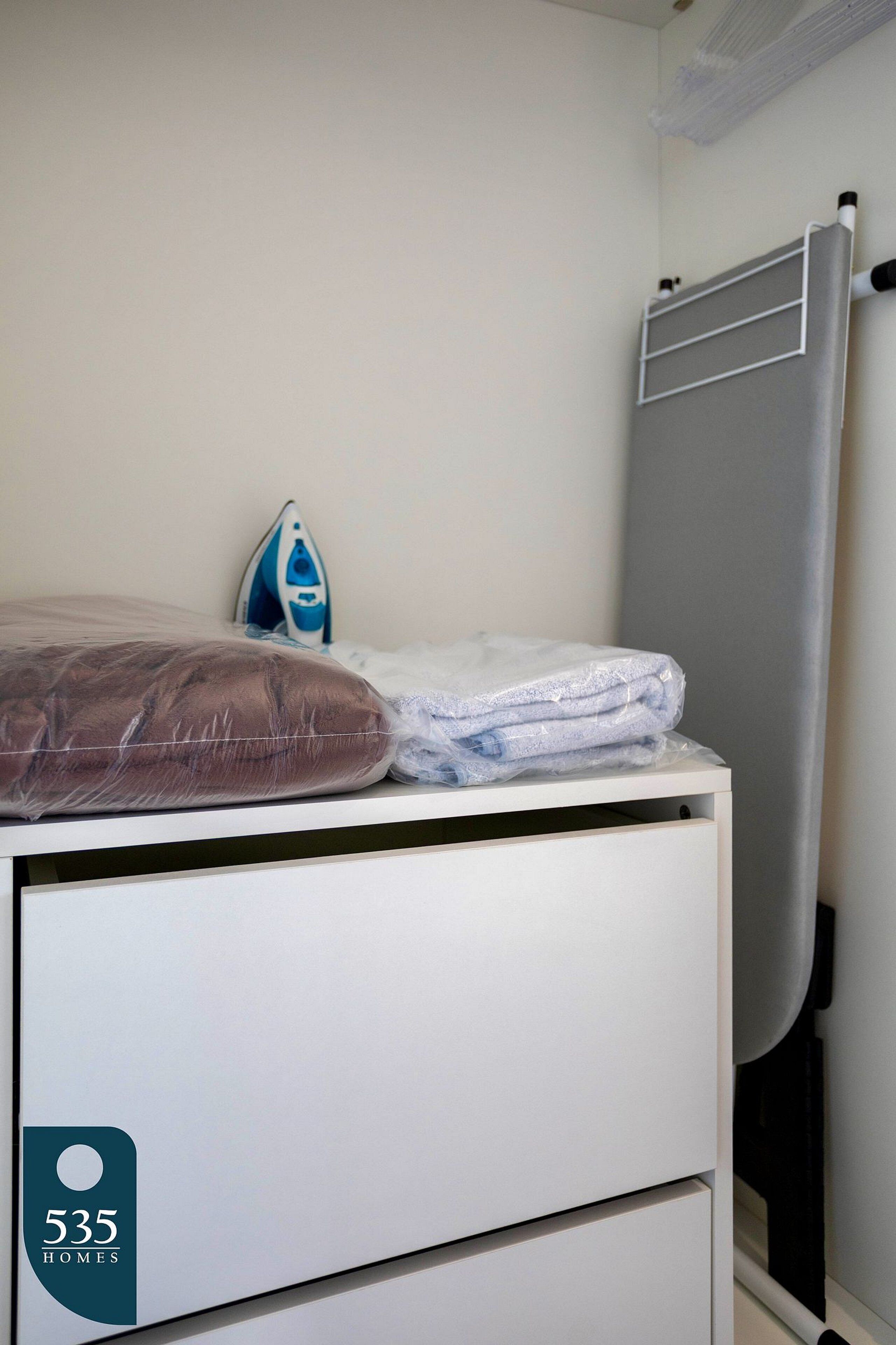 A neatly made bed with a pillow and folded towels, an iron, and a radiator in a room with a white wall and a door.