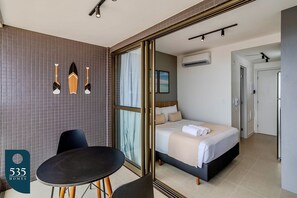 A modern hotel room with a bed, a dining table, and chairs. There are wall-mounted lights and a window with curtains. - Ocean View Studio on the Rio Vermelho Waterfront (Salvador)