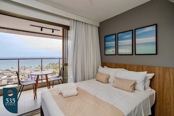A hotel room with a large bed, a round table with chairs, a balcony with a view of the city, and three framed pictures on the wall. - Ocean View Studio on the Rio Vermelho Waterfront (Salvador)