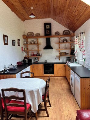 A compact kitchen with wooden cabinets, a central island, a dining table with chairs, and a washing machine. - Rural Donegal Cottage Near Sandy Beaches (Letter)