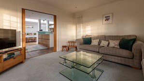 Ein modernes Wohnzimmer mit einem Glastisch, einer Couch mit Zierkissen, einem Fernseher auf einem Holzständer und Blick in den Küchenbereich.