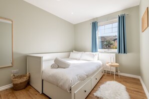Ein Schlafzimmer mit Bett, Fenster mit Vorhängen, Heizkörper, einem kleinen Tisch und einem Korb.