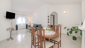 Zona pranzo con tavolo rotondo, sedie, televisore e divano in una stanza luminosa e bianca con pavimento piastrellato.