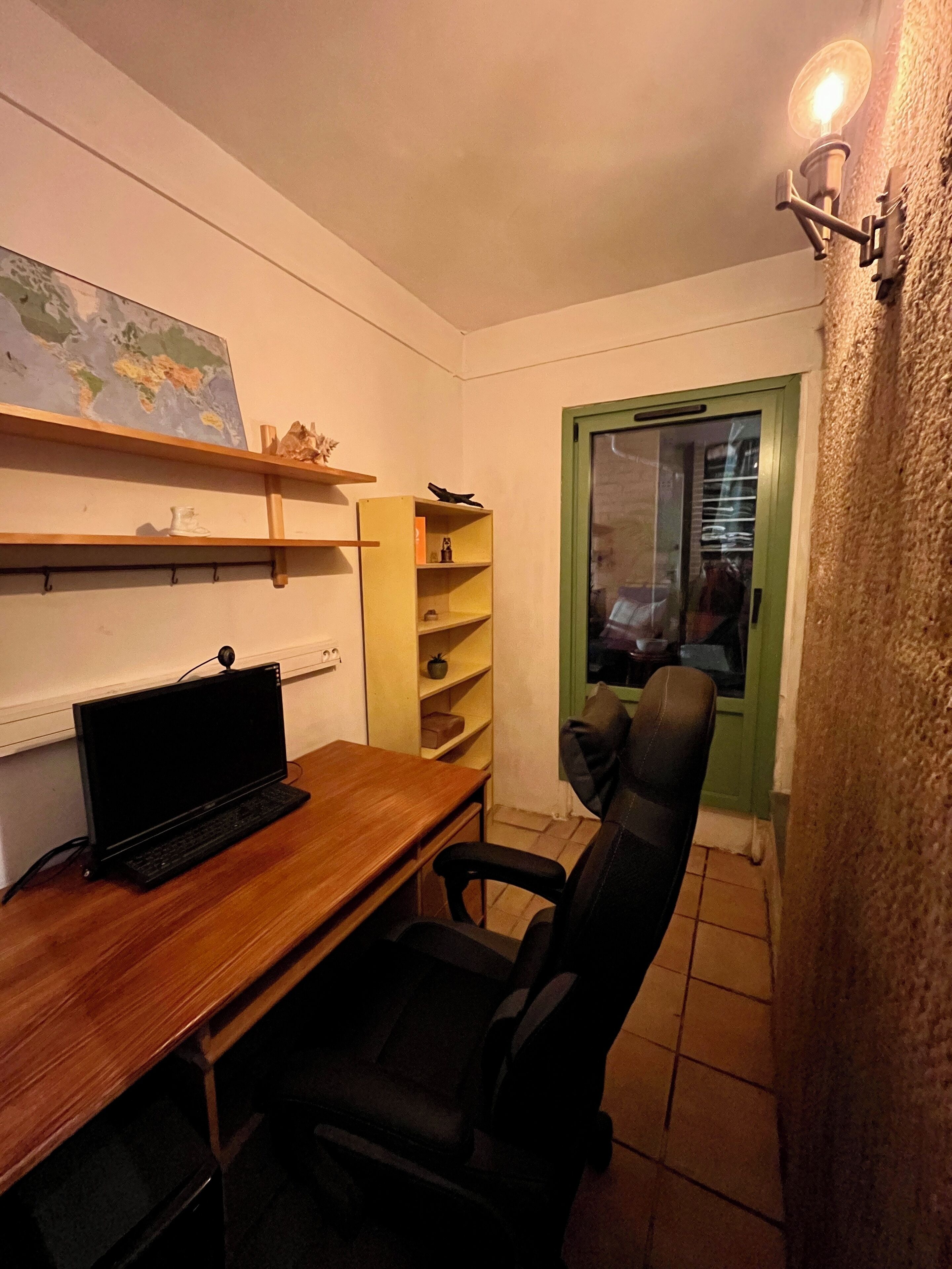 Une petite pièce faiblement éclairée, avec un bureau en bois, un fauteuil de bureau noir, une carte accrochée au mur et une fenêtre encadrée de vert.
