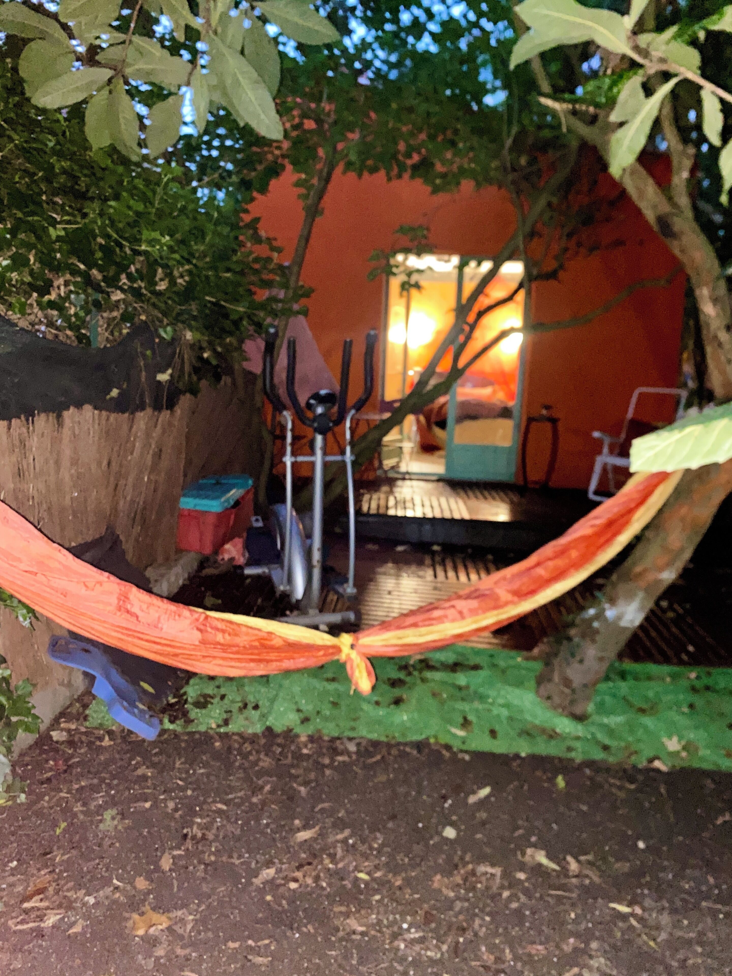 Un hamac est suspendu entre deux arbres dans un coin du jardin, à proximité d’une chaise et d’une table en métal.