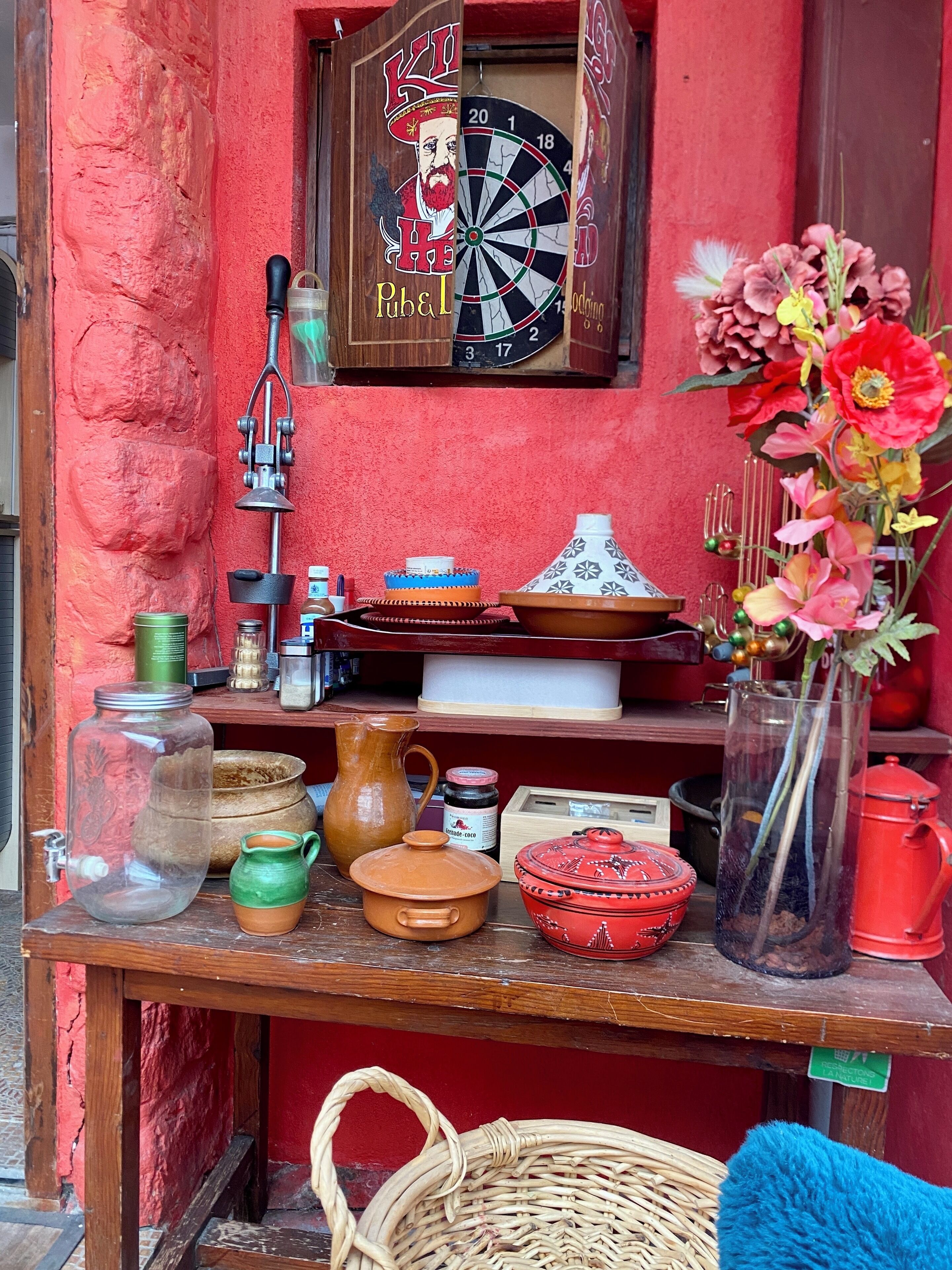 Un intérieur rustique avec un jeu de fléchettes, une étagère remplie de différents pots et récipients, et un vase avec des fleurs.
