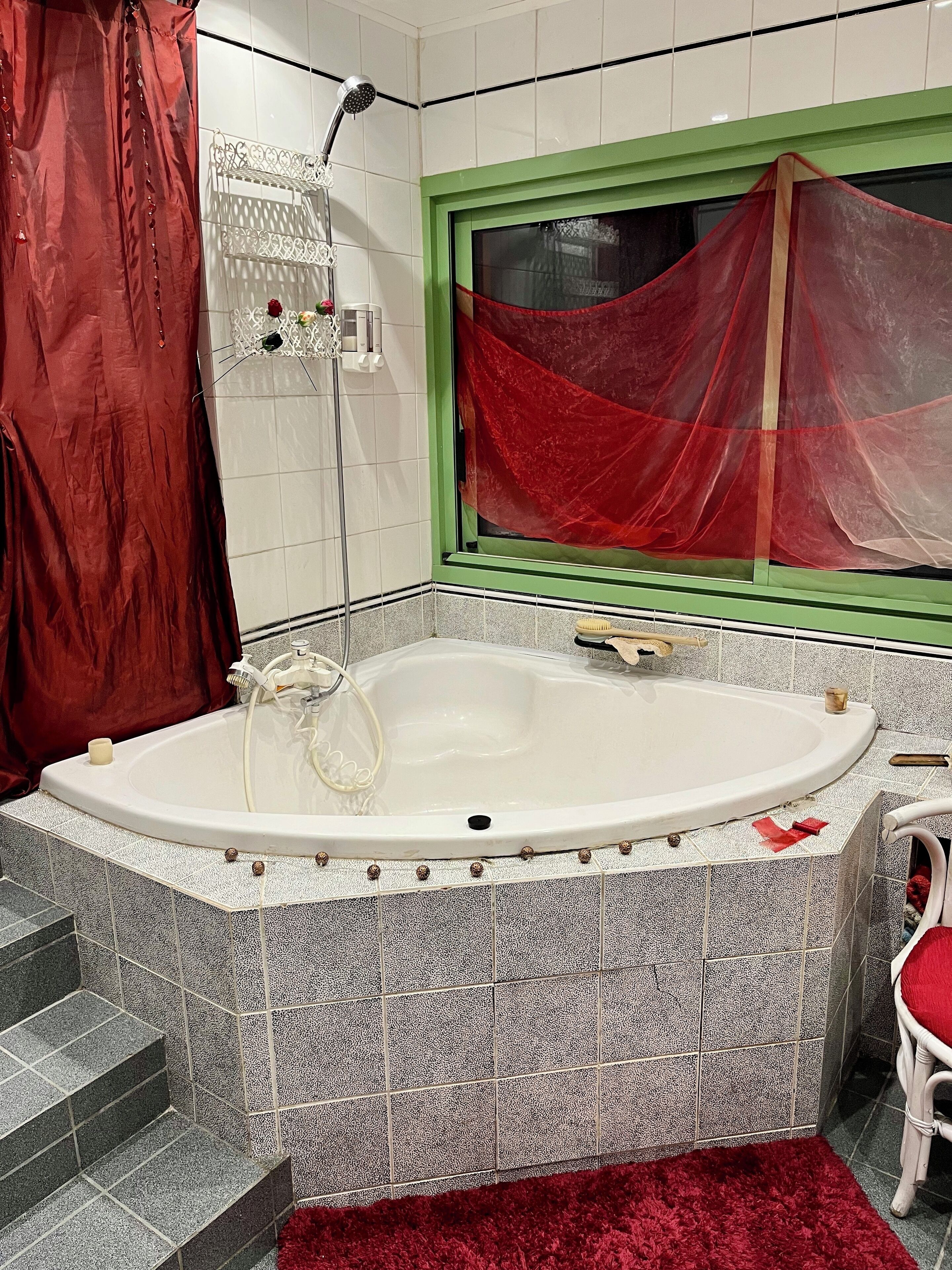 Een badkamer met een vrijstaand bad, een douchekop, een raam met rood gordijn en een rood vloerkleed.