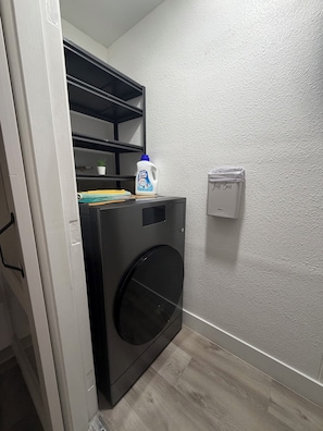 A compact laundry room with a front-loading washing machine, a shelf with folded clothes, a bottle of detergent, and a wall-mounted laundry basket. - Cozy 1-bedroom apartment in beautiful Las Vegas with AC (Las Vegas)