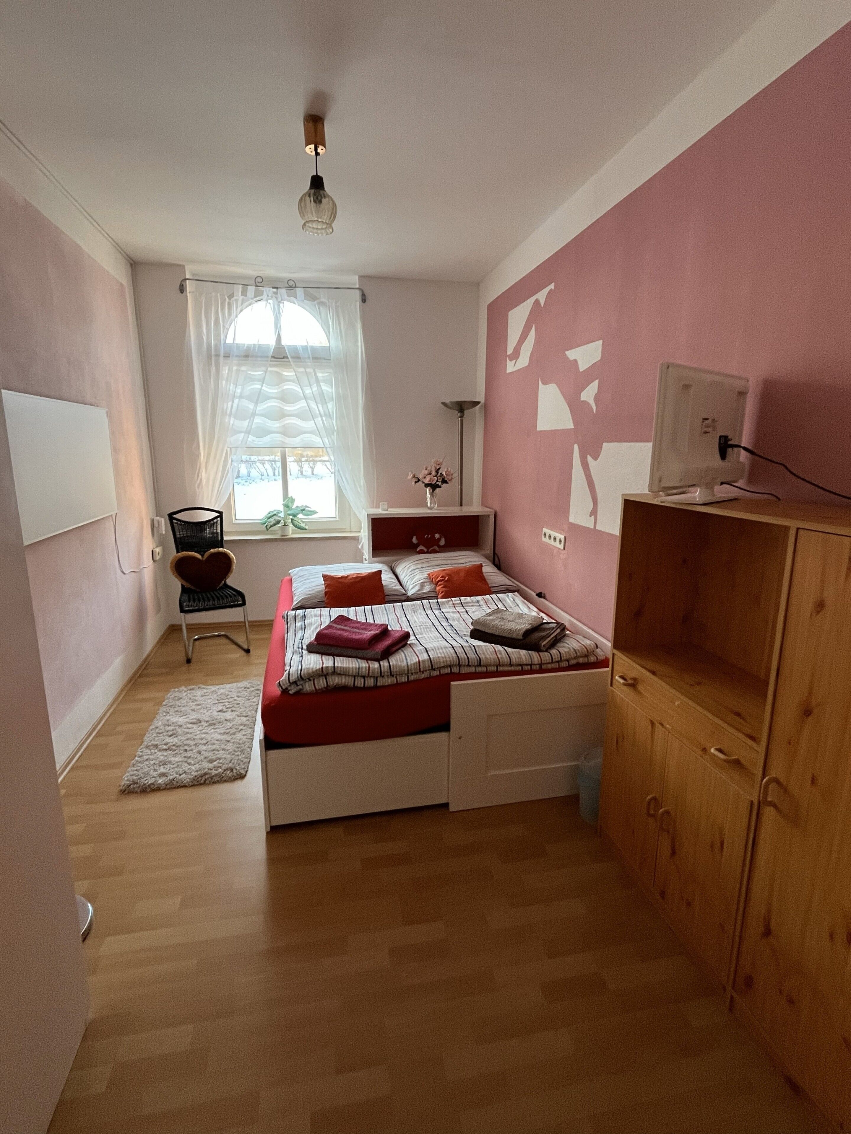 Een slaapkamer met een bed, een bureau, een stoel, een kledingkast, een raam met gordijnen en een plafondlamp.