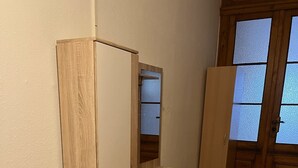 A room with a wooden wardrobe, a small cabinet, a black mat on the floor, and a wooden door with frosted glass panels.