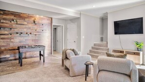 A modern living room with a wooden accent wall, a foosball table, a grey sofa set, a flat-screen TV, and a staircase. - Clemens House - Relaxing Lexington Home for Long Stays (Lexington)