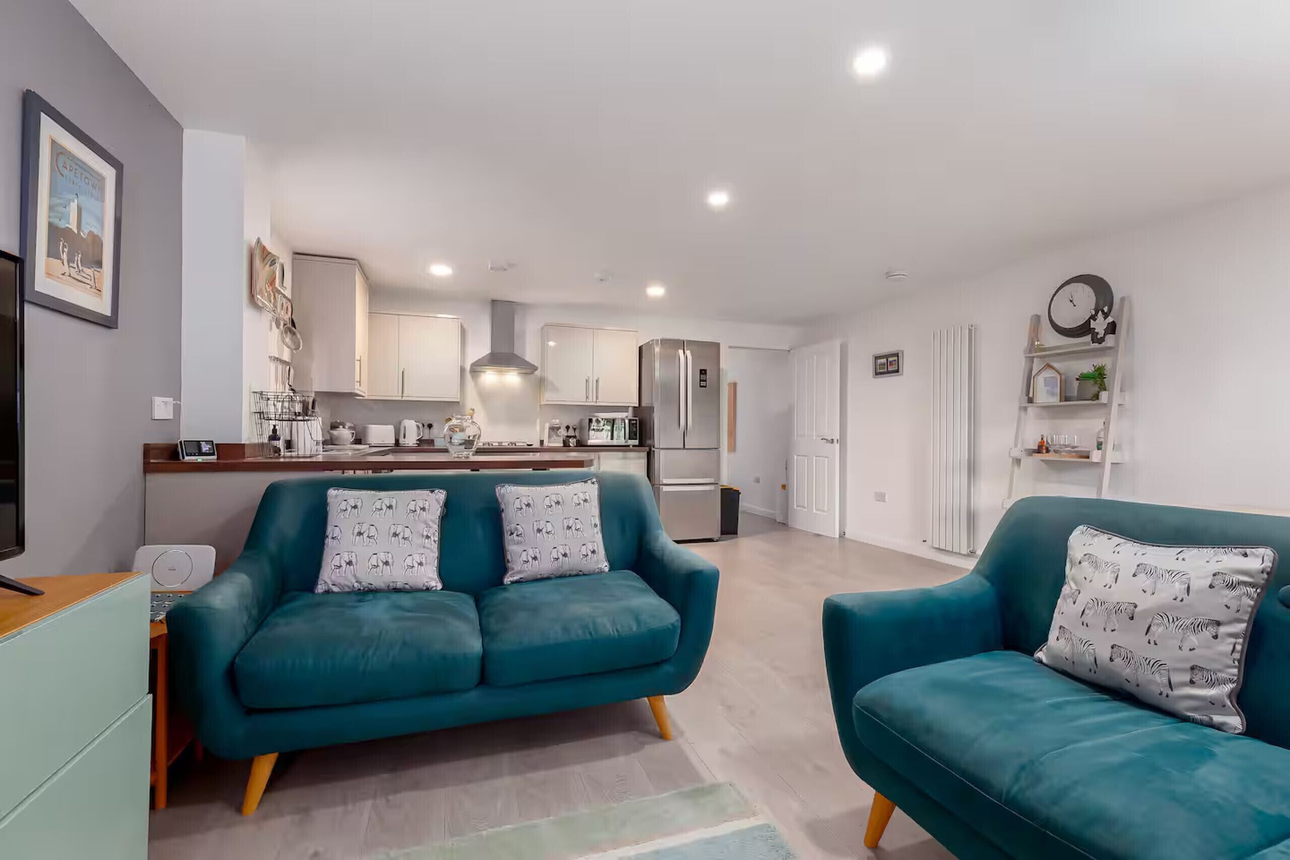 A modern living room with teal sofas, a kitchen in the background, and a white door leading to another room.