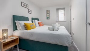 Una cama bien hecha con ropa blanca y un cabecero azul verdoso, una mesita de noche con lámpara y una ventana con persianas.