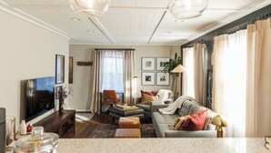 Ein modernes Wohnzimmer mit Sofa, Couchtisch und Fernseher. Vorhänge, eine Pflanze und gerahmte Bilder sind an der Wand zu sehen.