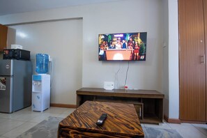 A living room with a flat-screen TV mounted on the wall, a wooden coffee table with a remote control, a refrigerator, a water dispenser, and a wooden cabinet. - Charming apartment in Nairobi near JKIA/SGR perfect for your next getaway (Nairobi)