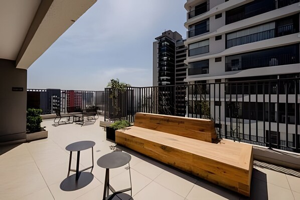 Eine Dachterrasse mit einer Holzbank, zwei runden Tischen und Blick auf ein modernes Gebäude.
