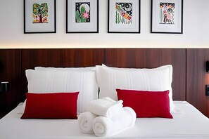 Ein ordentlich bezogenes Bett mit weißen und roten Kissen, ein aufgerolltes weißes Handtuch und vier gerahmte Kunstwerke an der Wand darüber.