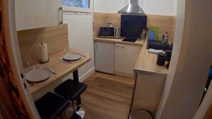 A compact kitchen with a microwave, stove, and sink. There is a window above the sink and a small trash bin near the door.