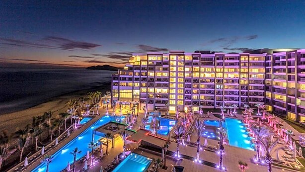 Un hotel de varios pisos con zona de piscina, rodeado de palmeras y vistas a la playa.