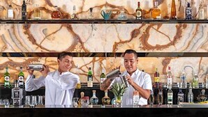 Un bar con dos cocteleros preparando bebidas. El bar tiene una pared que simula mármol y estantes con diferentes botellas. Hay dos taburetes de metal frente a la barra.