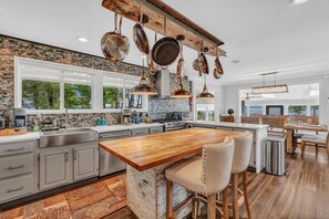 A modern kitchen with a central island, hanging pots, and a dining area with wooden chairs. -  Oceanfront Seaglass Estate  Near Acadia National Park, with Hot Tub and Views! (Gouldsboro)