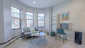 A modern living room with a grey sofa, a round coffee table, and a blue rug. There are three windows with blinds, a bookshelf, and a painting on the wall. - Blissful South End Studio (Boston)