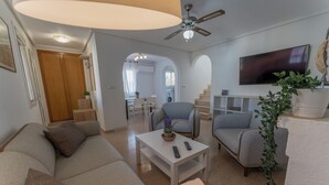 Un salon avec un canapé, des fauteuils, une table basse et un téléviseur. On aperçoit au fond un ventilateur de plafond, un escalier et une cuisine.