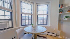 Een hoekkamer met twee ramen, een ronde houten tafel, twee stoelen, een radiator en een plank met boeken en planten.
