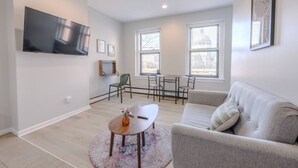 A modern living room with a grey sofa, a coffee table, a TV mounted on the wall, and a dining area with a table and chairs. - Chic South End Studio | Perfect for Nurses! (Boston)