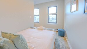 A bedroom with a bed, two windows, a rug, and a blue ottoman. - Chic South End Studio | Perfect for Nurses! (Boston)