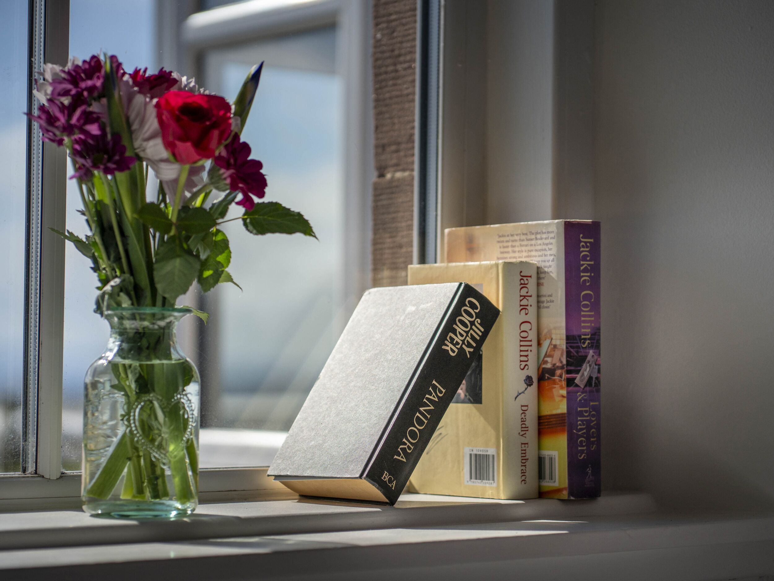 A window sill with a clear vase containing flowers, a book titled 'PANDORA', and other books.